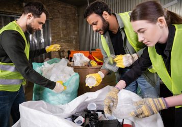 Indian sorter in high visibility vest and gloves holding plastic trash for recycling near sack while working with colleagues together in waste disposal station, garbage sorting and recycling concept